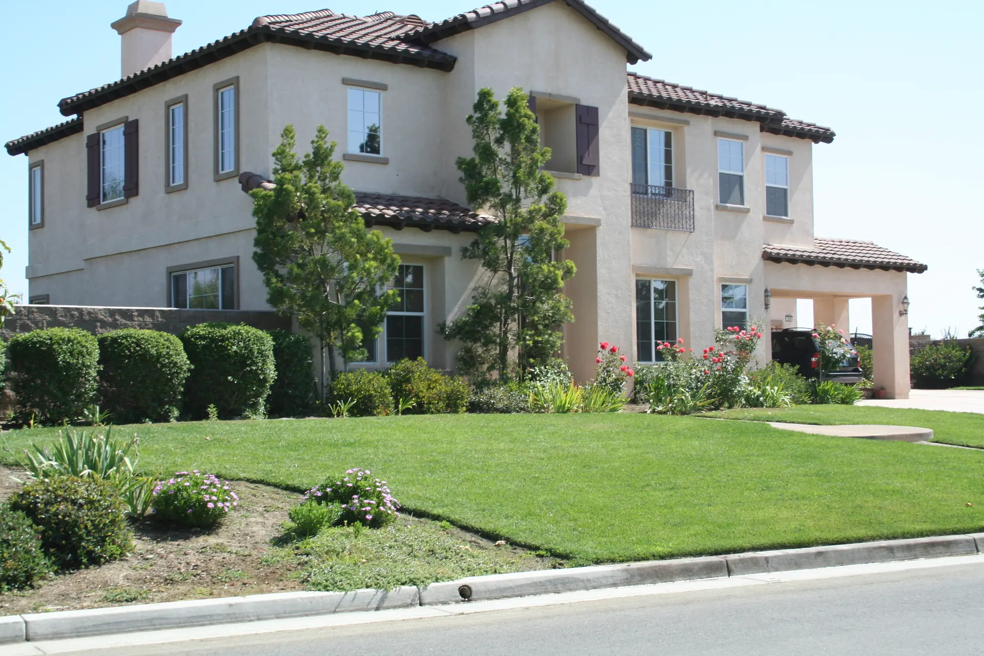 Two-story cream stucco house with a tile roof and dark shutters behind a sloping green lawn, rose bushes and trimmed shrubs, with a car parked under a covered porte-cochere at the right.