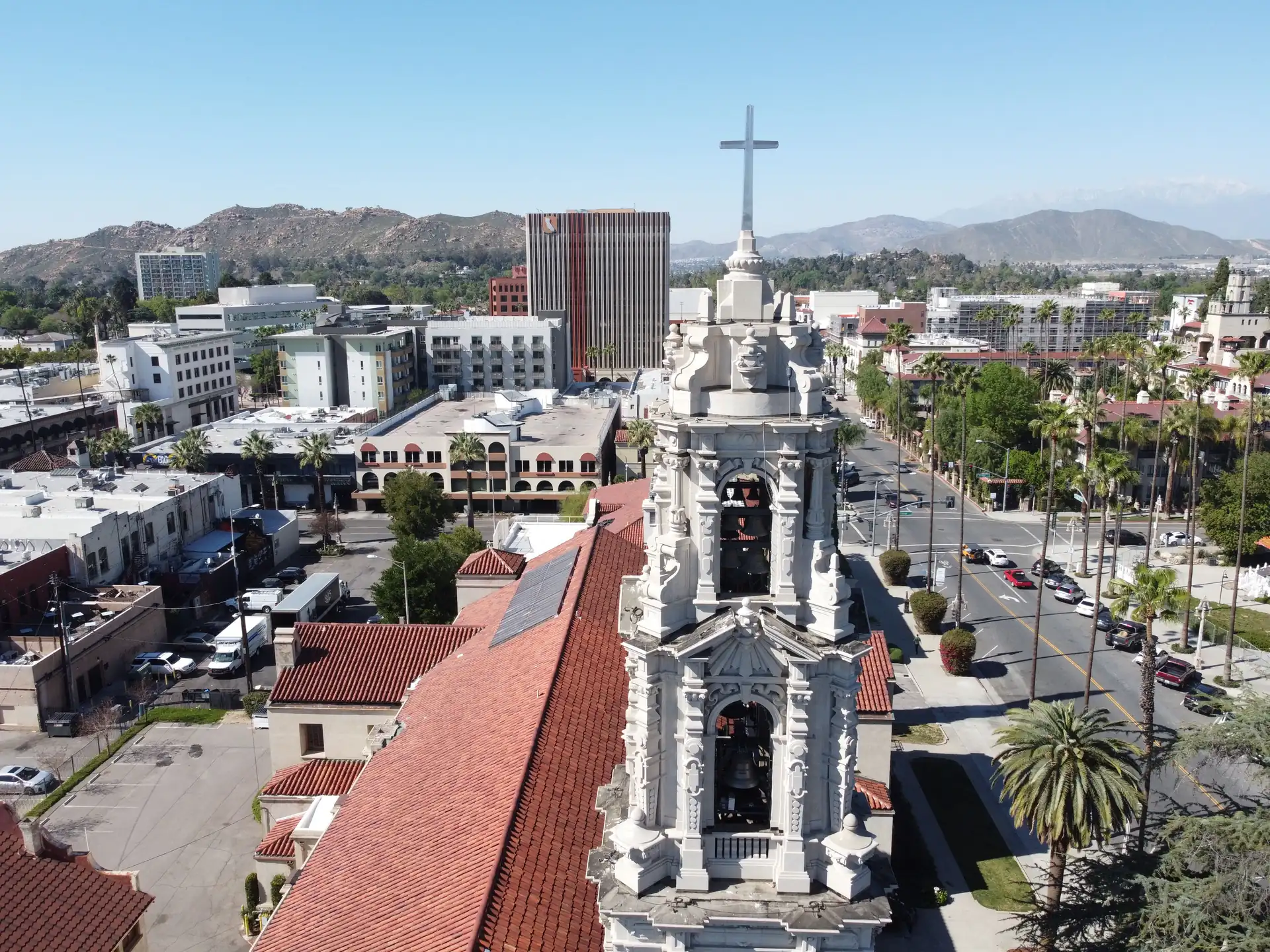 Aerial view of downtown Riverside, California, with the Mission Inn's red tile roof, a white bell tower, palm-lined streets, and the hills beyond.